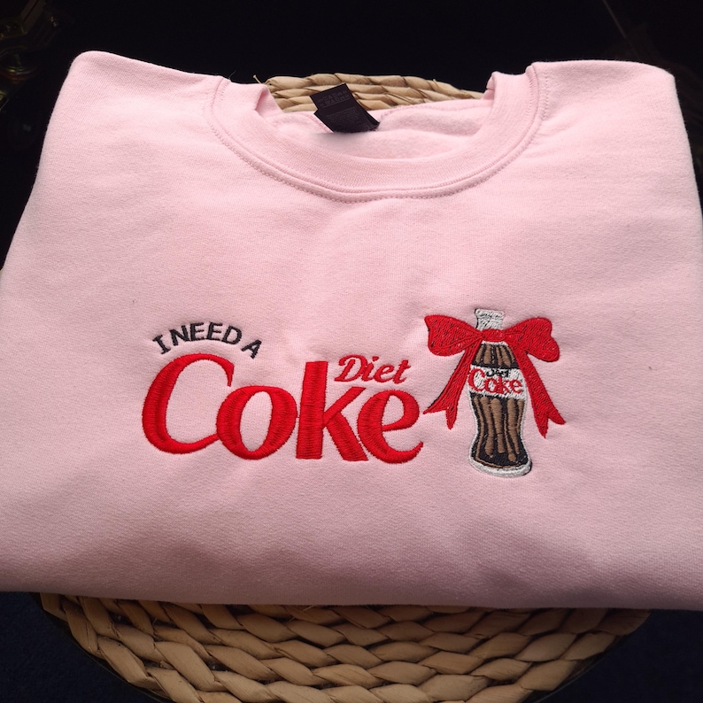 May include: A light pink sweatshirt with the embroidered phrase "I NEED A Diet Coke" in red. A Diet Coke bottle with a red bow is embroidered on the right side. The sweatshirt is folded on a woven surface.