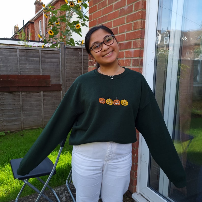 Puede incluir: Una sudadera verde oscuro con una fila de cuatro caras de calabaza talladas bordadas en naranja y amarillo. La sudadera la lleva puesta una persona con el pelo largo y oscuro y gafas. La persona est&aacute; de pie frente a una pared de ladrillo y una ventana.