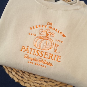 May include: Cream-colored sweatshirt with an orange embroidered design. The design features a pumpkin, a mouse, and the words "The Sleepy Hollow Pâtisserie" and "Pumpkin Pastries and Breads". The sweatshirt is folded on a woven basket.
