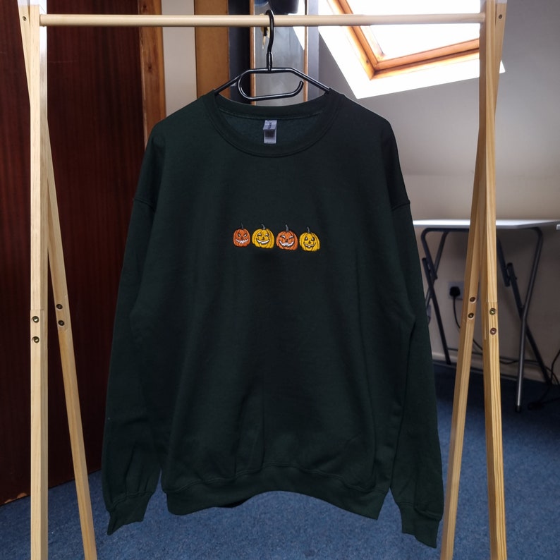 Puede incluir: Una sudadera verde oscuro con una fila de tres caras de calabaza talladas bordadas en naranja y amarillo.