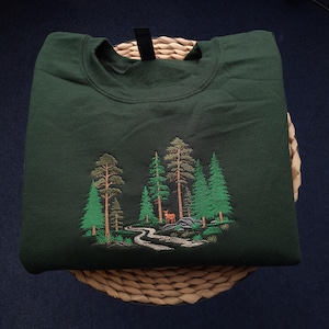 May include: Dark green sweatshirt featuring an embroidered forest scene. The design includes various green trees, a winding path, and a small deer. The sweatshirt is folded on a woven basket, with a black tag visible at the neckline.