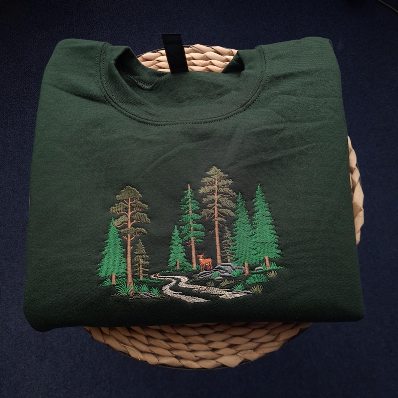 May include: Dark green sweatshirt featuring an embroidered forest scene. The design includes various green trees, a winding path, and a small deer. The sweatshirt is folded on a woven basket, with a black tag visible at the neckline.