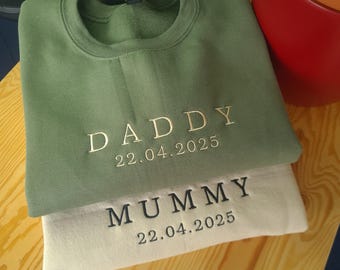May include: Two sweatshirts, one green and one cream, are stacked. The green sweatshirt has "DADDY" embroidered in gold thread, with the date "22.04.2025" below. The cream sweatshirt has "MUMMY" and the same date embroidered. The sweatshirts are on a wooden surface.