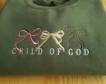 May include: A green sweatshirt with three embroidered bows in pink, gold, and green. The text "CHILD OF GOD" is embroidered in white below the bows.