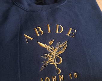 Può includere: Felpa blu navy con ricamo dorato che recita "ABIDE JOHN 16" e presenta un design di pianta stilizzato.