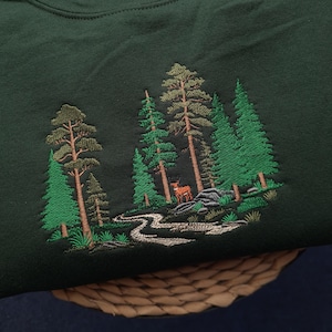 May include: Dark green sweatshirt with an embroidered forest scene. The design features tall green trees, a winding river, and a deer. The embroidery is in shades of green, brown, and white, creating a detailed nature-themed design.