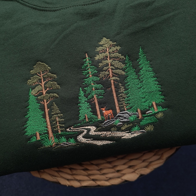 May include: Dark green sweatshirt with an embroidered forest scene. The design features tall green trees, a winding river, and a deer. The embroidery is in shades of green, brown, and white, creating a detailed nature-themed design.