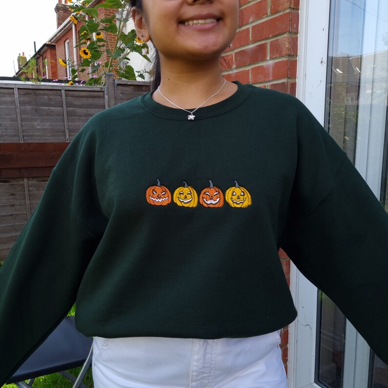 Puede incluir: Una sudadera verde oscuro con una fila de cuatro caras de calabaza talladas bordadas en naranja y amarillo.