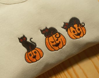 Puede incluir: Sudadera beige con diseños bordados de Halloween. Tres calabazas naranjas con caras sonrientes están coronadas por un gato negro. Los gatos tienen ojos rojos. Una prenda acogedora y festiva para la temporada de miedo.