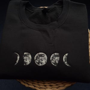 May include: Black sweatshirt featuring an embroidered design of the phases of the moon in white. The design includes crescent, half, and full moon shapes. The sweatshirt is made of a soft material and has a crew neck.