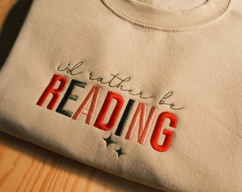 Puede incluir: Una sudadera beige con el texto "I'd rather be READING" bordado en rojo, rosa, negro y verde. El texto está en una fuente en negrita y en bloques.