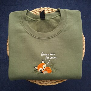 May include: Olive green sweatshirt with a crew neck. The sweatshirt features an embroidered sleeping fox with the text "Giving zero fox today". The sweatshirt is folded on a woven basket.