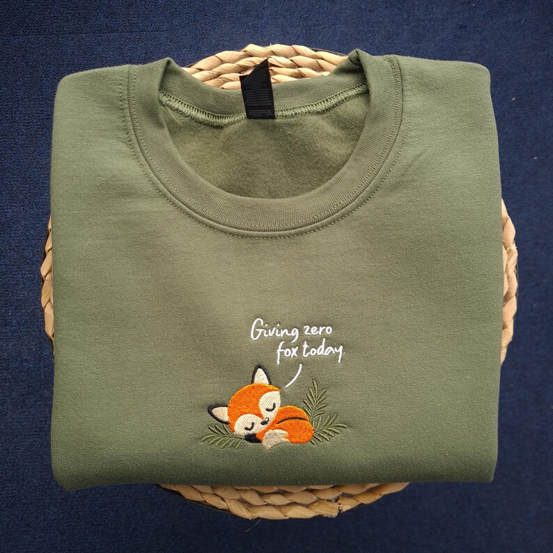 May include: Olive green sweatshirt with a crew neck. The sweatshirt features an embroidered sleeping fox with the text "Giving zero fox today". The sweatshirt is folded on a woven basket.