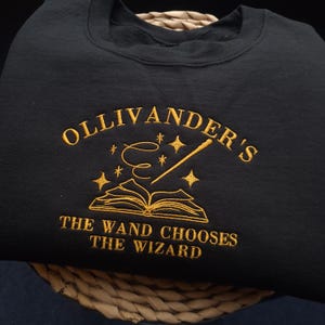 May include: A black crewneck sweatshirt featuring gold embroidery. The text reads OLLIVANDER'S and THE WAND CHOOSES THE WIZARD. Between the text is an embroidered open book, a magic wand, and several small four-pointed stars.