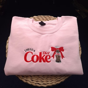 May include: A light pink sweatshirt with the embroidered phrase "I Need a Diet Coke" in red. A Diet Coke bottle with a red bow is embroidered on the right side. The sweatshirt is folded on a woven basket.