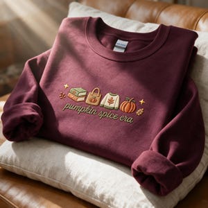 Puede incluir: Una sudadera de cuello redondo color granate con un diseño bordado de libros apilados, una bolsa de tela, un suéter y una calabaza. El texto "pumpkin spice era" está bordado en cursiva debajo de los iconos, junto a estrellas y hojas.