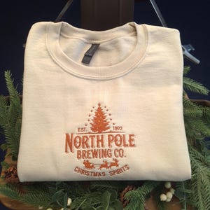 May include: A beige sweatshirt featuring embroidered text and a festive design. The text reads "North Pole Brewing Co." with a Christmas tree and "Christmas Spirits" in orange thread. The folded sweatshirt is displayed on a wooden surface.