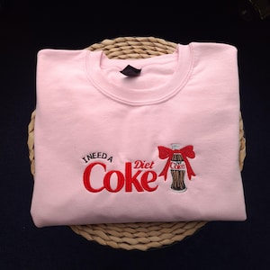 May include: A light pink sweatshirt with the embroidered phrase "I NEED A Diet Coke" in red. A Diet Coke bottle with a red bow is embroidered on the right side. The sweatshirt is folded on a woven mat.