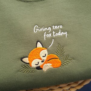 May include: Olive green sweatshirt with an embroidered sleeping fox. The fox is orange and white with black accents, nestled among green leaves. The text "Giving zero fox today" is written above the fox in white script.