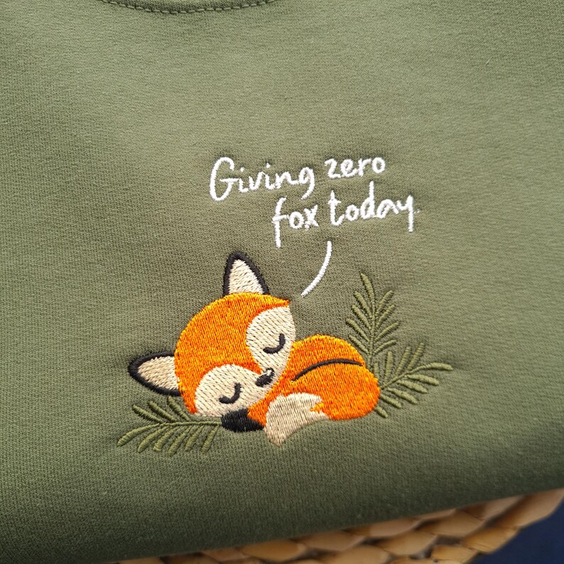 May include: Olive green sweatshirt with an embroidered sleeping fox. The fox is orange and white with black accents, nestled among green leaves. The text "Giving zero fox today" is written above the fox in white script.