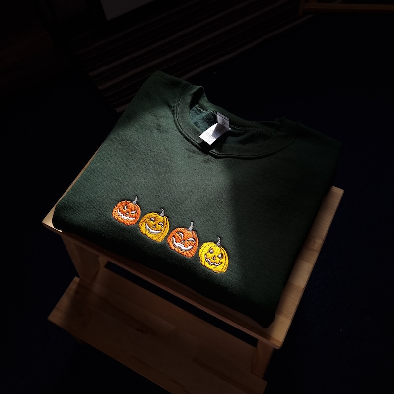 Puede incluir: Una sudadera verde oscuro con un dise&ntilde;o bordado de cuatro calabazas sonrientes. Las calabazas son de color naranja y amarillo con ojos y bocas negras.
