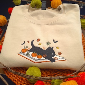 Può includere: Felpa color crema con un ricamo di un gatto nero sdraiato su un libro aperto con zucche e foglie autunnali. Il design include un pipistrello e altri elementi a tema autunnale.