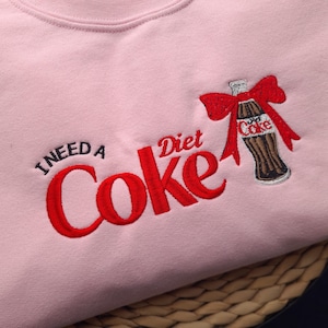 May include: A light pink sweatshirt with the embroidered phrase "I NEED A Coke Diet" in red thread. A Diet Coke bottle with a red bow is embroidered on the right side. The sweatshirt is a casual, comfortable style.