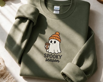 Op de afbeelding: Een olijfgroene sweater met ronde hals en een geborduurde afbeelding van een witte geest met een oranje gebreide muts die een koffiebeker met een pompoenpictogram vasthoudt. De tekst onder de geest luidt SPOOKY season in zwarte letters.