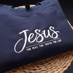 May include: A navy blue sweatshirt featuring the word "Jesus" in white script embroidery. Beneath, the phrase "THE WAY THE TRUTH THE LIFE" is displayed in white capital letters. The garment is made from a soft fabric.
