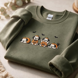 Peut inclure: Un sweat-shirt à col rond vert olive orné d'une broderie représentant quatre petits chiots vêtus de costumes de citrouille orange. Les chiots sont représentés dans diverses postures, accompagnés de petites silhouettes de chauves-souris noires.