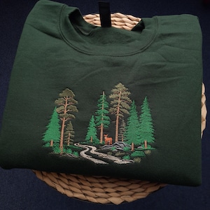 May include: Dark green sweatshirt with an embroidered forest scene. The design features green trees, a winding path, and a deer. The sweatshirt is folded on a woven basket.