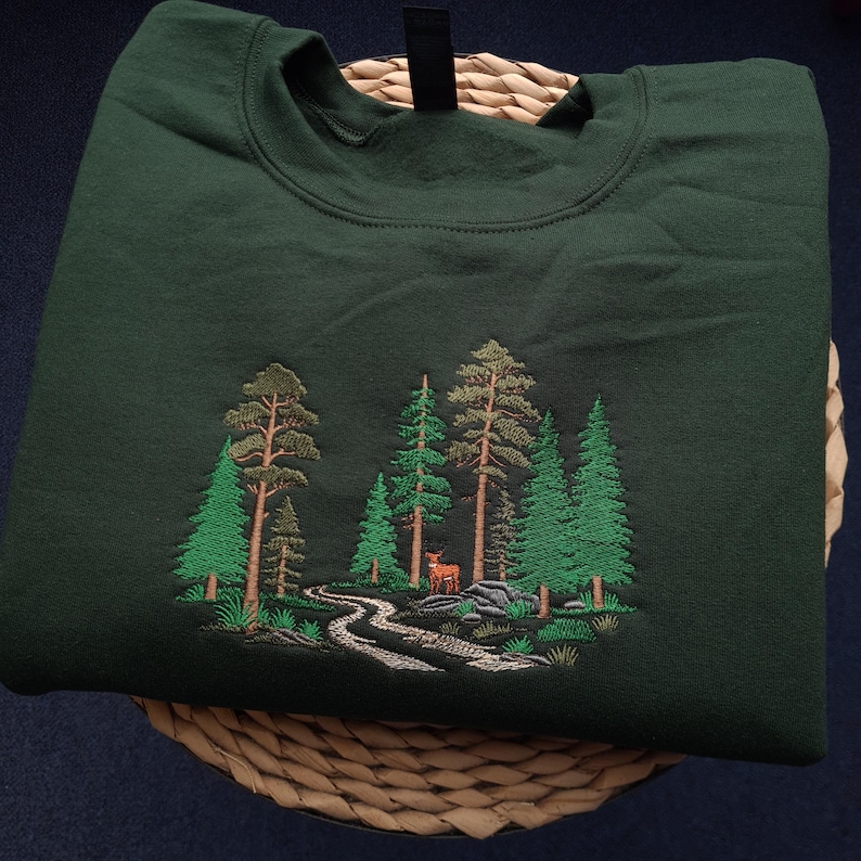 May include: Dark green sweatshirt with an embroidered forest scene. The design features green trees, a winding path, and a deer. The sweatshirt is folded on a woven basket.
