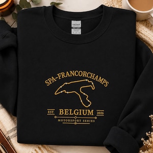 Puede incluir: Una sudadera negra de manga larga con bordado dorado en el pecho. El diseño muestra el mapa del circuito de Spa-Francorchamps, el texto "SPA-FRANCORCHAMPS", "EST. BELGIUM 2026" y "MOTORSPORT SERIES" con detalles de líneas decorativas.