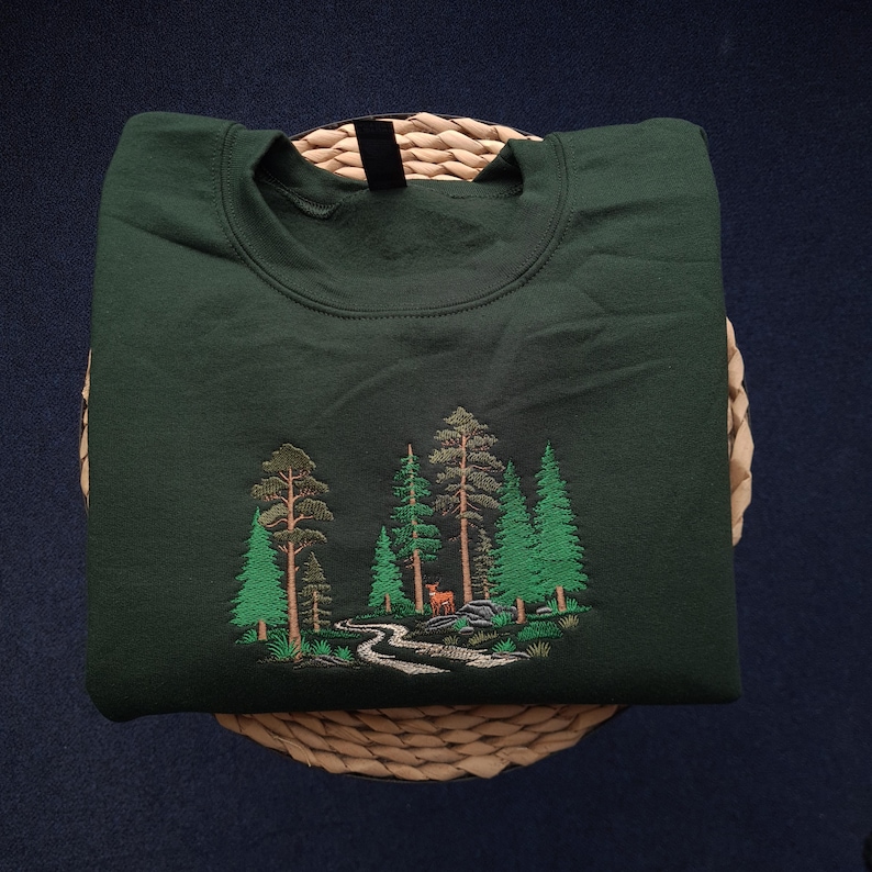 May include: Dark green sweatshirt with an embroidered forest scene. The design features various green trees, a winding river, and a small deer. The sweatshirt is folded neatly on a woven basket, set against a dark blue background.
