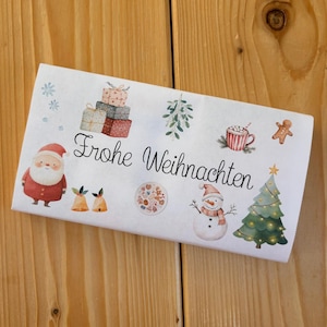 May include: A rectangular white paper napkin featuring the black cursive text Frohe Weihnachten surrounded by colorful illustrations of a Santa Claus, stacked gift boxes, mistletoe, a mug, a gingerbread man, bells, cookies, a snowman, and a tree.