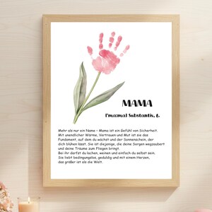 Peut inclure: Un cadre en bois clair encadre une impression avec une empreinte de main rose en forme de fleur. Le mot "MAMA" est écrit en noir. Un texte en allemand est affiché en dessous.