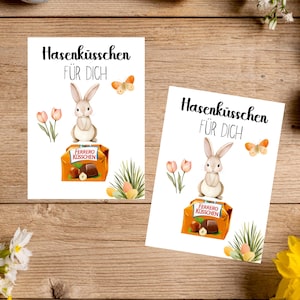 Peut inclure: Deux cartes de vœux de Pâques avec le texte "Hasenküsschen FÜR DICH" et des illustrations d'un lapin, de papillons et d'une boîte de chocolats. Les cartes sont entourées d'œufs de Pâques, de fleurs et d'éléments décoratifs sur une surface en bois.