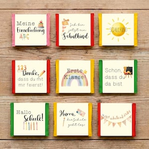 Puede incluir: Nueve pequeñas barras de chocolate envueltas con etiquetas coloridas y texto en alemán. Las etiquetas presentan frases como "Meine Einschulung" y "Erste Klasse!" con ilustraciones. Dispuestas sobre una superficie de madera.