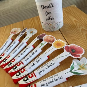 May include: A selection of gifts featuring floral designs. The image shows decorated candy bars with flower-shaped toppers and inspirational messages. A white paper cup with the words "Danke f&uuml;r alles" and a floral border is also present.