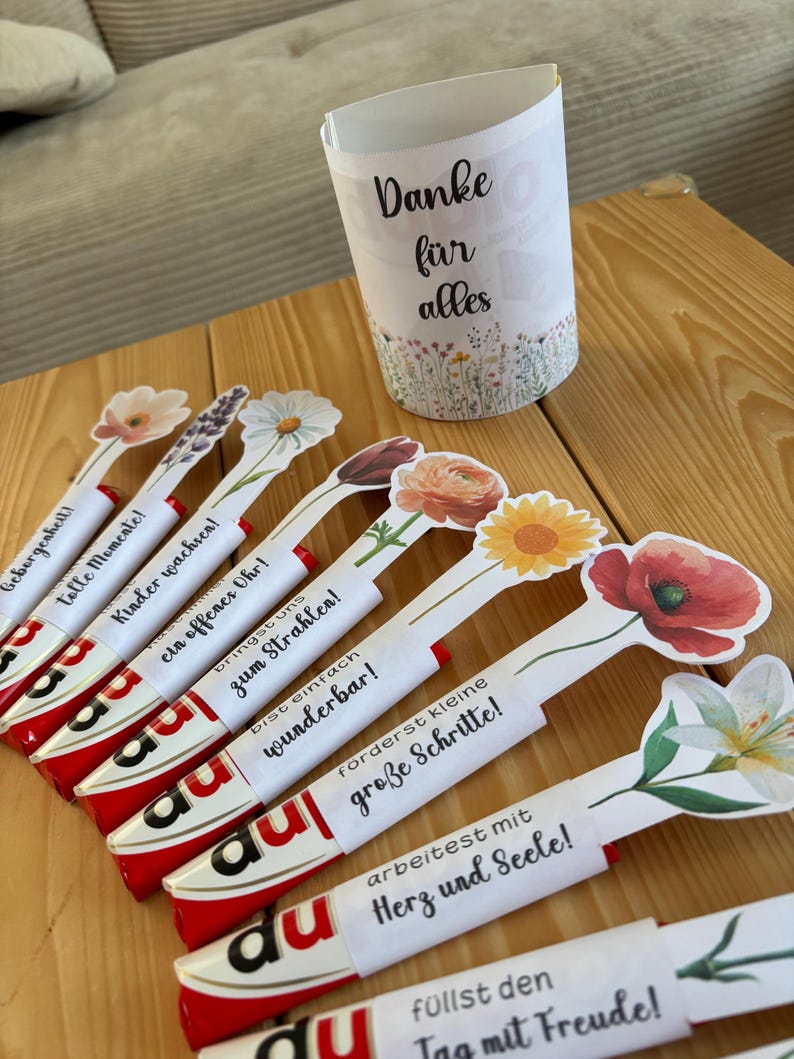 May include: A selection of gifts featuring floral designs. The image shows decorated candy bars with flower-shaped toppers and inspirational messages. A white paper cup with the words "Danke f&uuml;r alles" and a floral border is also present.