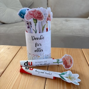 May include: A white paper cup with the text "Danke f&uuml;r alles" holds several paper flower cutouts. Two decorative paper sticks with flower cutouts and German text are on a wooden surface.