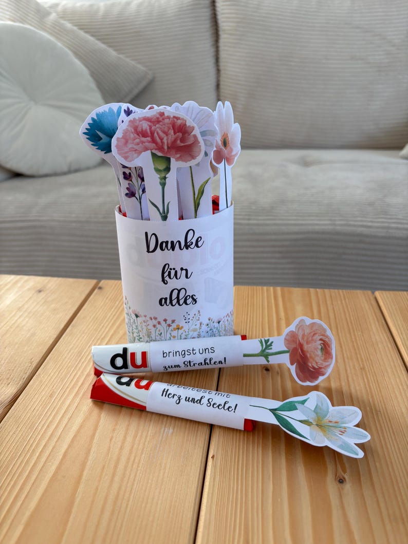 May include: A white paper cup with the text "Danke f&uuml;r alles" holds several paper flower cutouts. Two decorative paper sticks with flower cutouts and German text are on a wooden surface.
