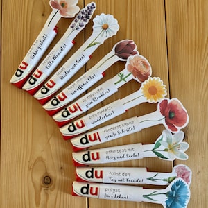 May include: A selection of candy bar wrappers with floral decorations and German text. Each wrapper has a different flower and a positive message. The wrappers are displayed on a wooden surface.