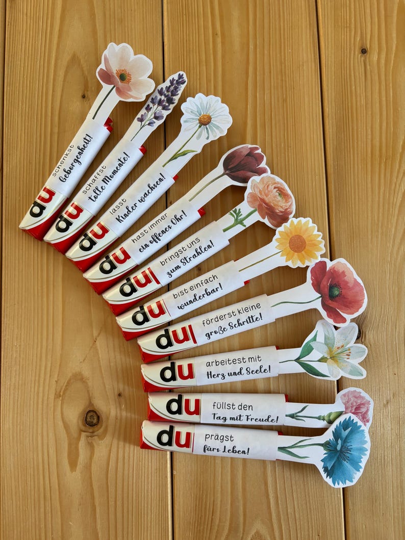 May include: A selection of candy bar wrappers with floral decorations and German text. Each wrapper has a different flower and a positive message. The wrappers are displayed on a wooden surface.