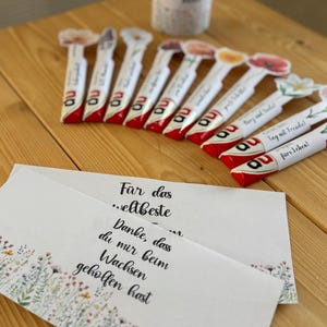 May include: A selection of white and red wrapped candies with floral designs and the letters "au" printed on them. Several cards with floral borders and German text are also visible, alongside a white cylindrical container with text.