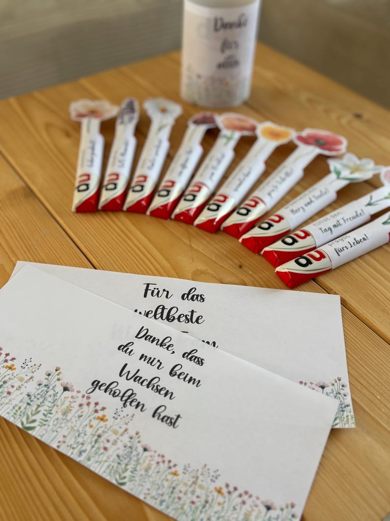 May include: A selection of white and red wrapped candies with floral designs and the letters "au" printed on them. Several cards with floral borders and German text are also visible, alongside a white cylindrical container with text.