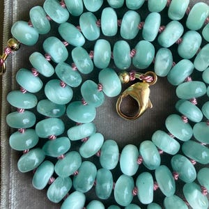 May include: A necklace featuring smooth, round, light mint green beads strung on a thin pink cord. The beads have a translucent, polished finish. The necklace is secured with a gold-toned metal lobster claw clasp and a small gold-toned bead.