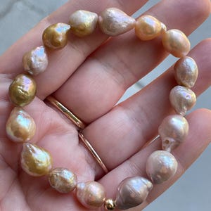 May include: A bracelet with irregularly shaped, iridescent pearls in gold, pink, and cream hues. The pearls are strung together with a gold-coloured bead clasp. This is a piece of jewellery.