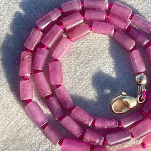 Può includere: Una collana con perline cilindriche rosa dalla consistenza marmorizzata, infilate su un cordoncino rosa acceso. La collana include piccole perline distanziatrici rosa tra i cilindri ed è rifinita con una chiusura a moschettone dorata.