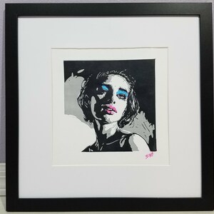May include: Framed black and white print of a woman with blue eyeshadow and pink lipstick. The artwork is in a white mat and black frame. The woman's portrait is a striking contrast of light and shadow.